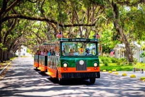 A green and orange trolley is driving down a tree lined street while offering charter flights in Florida.