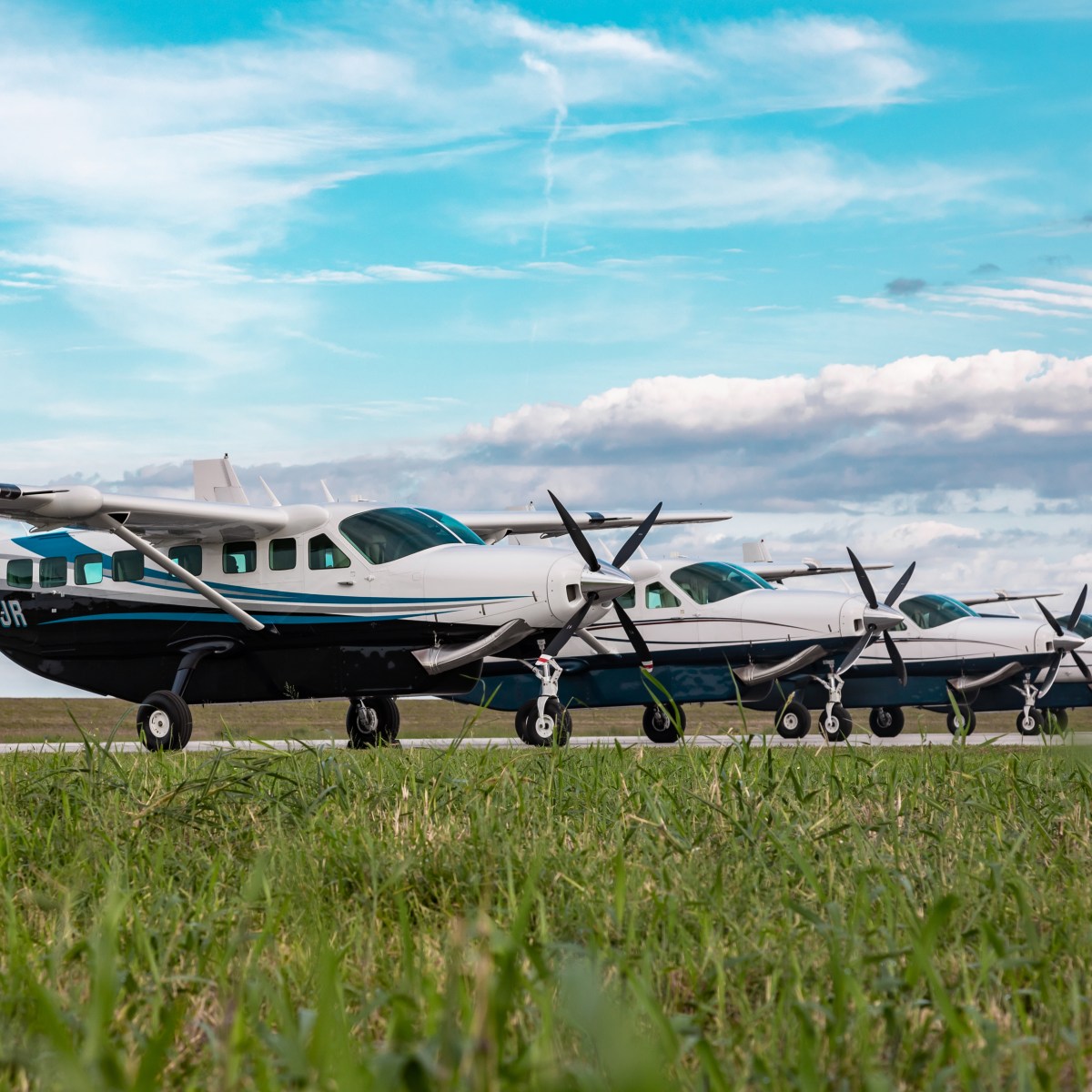 Four small aircraft parked on a grassy field under a cloudy blue sky.