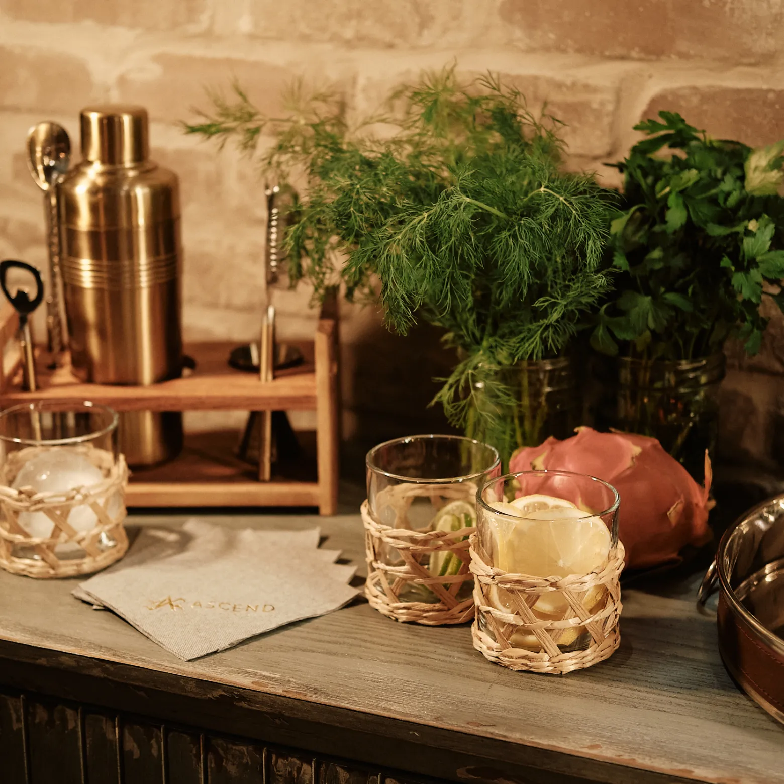 Cocktail setup with glasses, shaker, herbs, and a liquor bottle on a wooden surface.