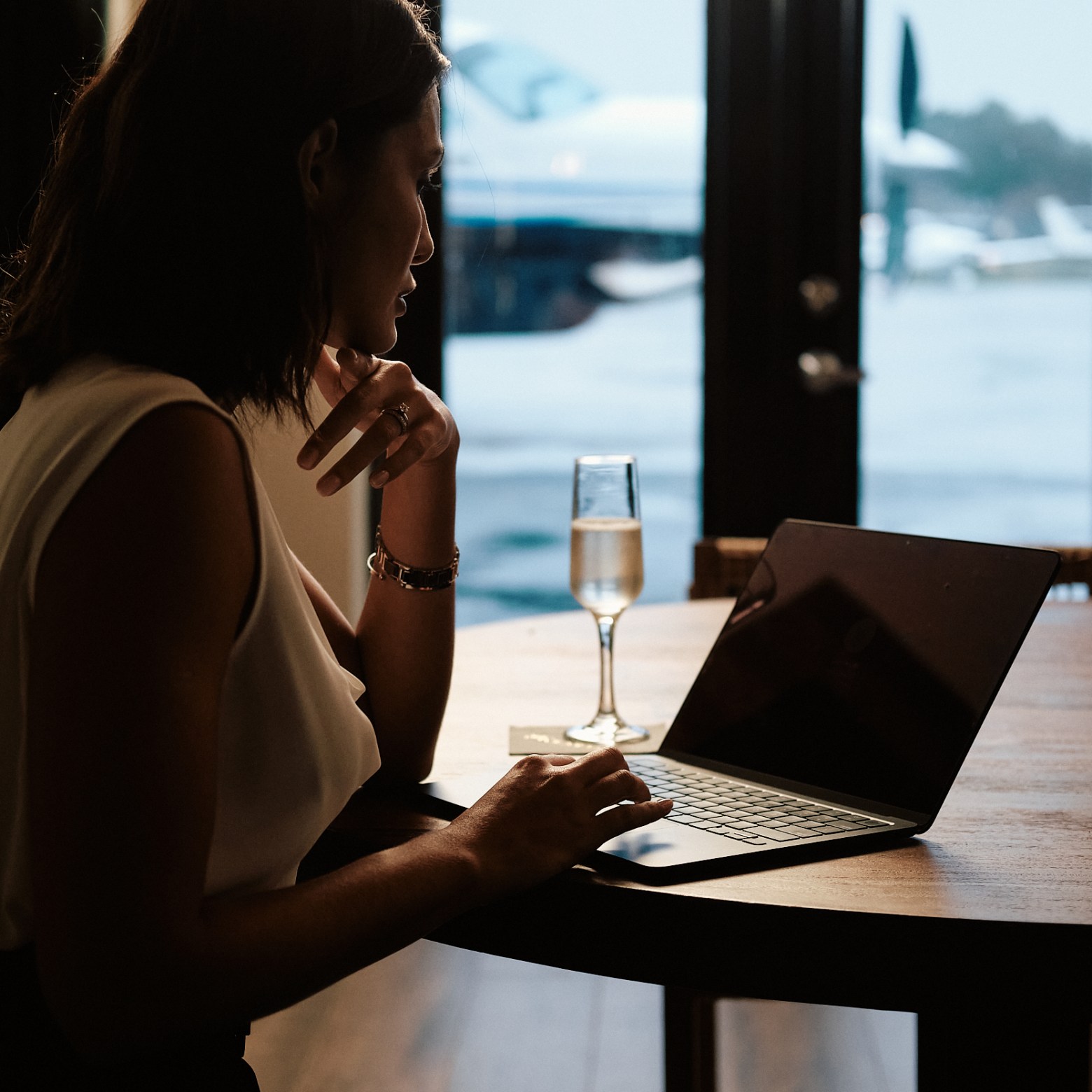 Woman using a laptop with a glass of champagne beside her, airplane visible through window.