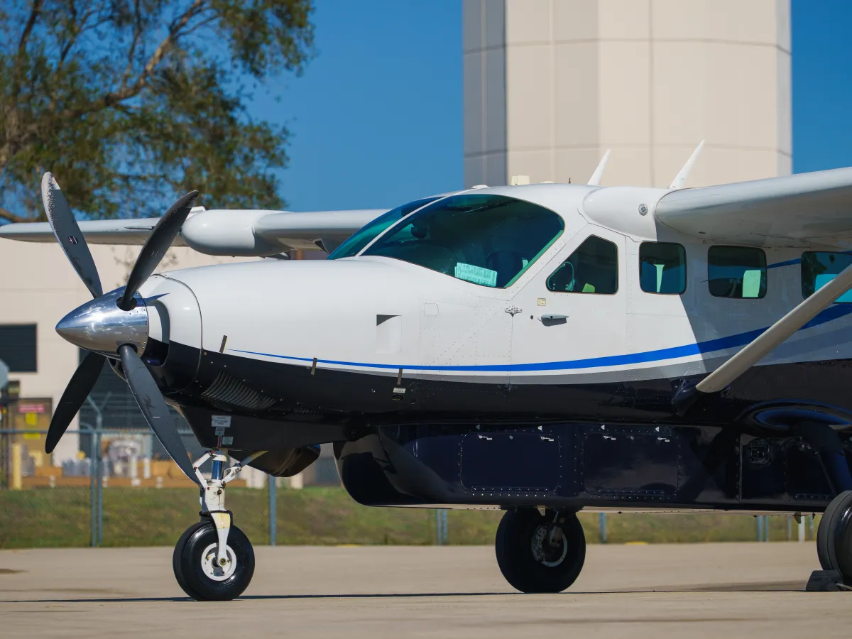 White and blue propeller plane on tarmac with control tower and trees in background.